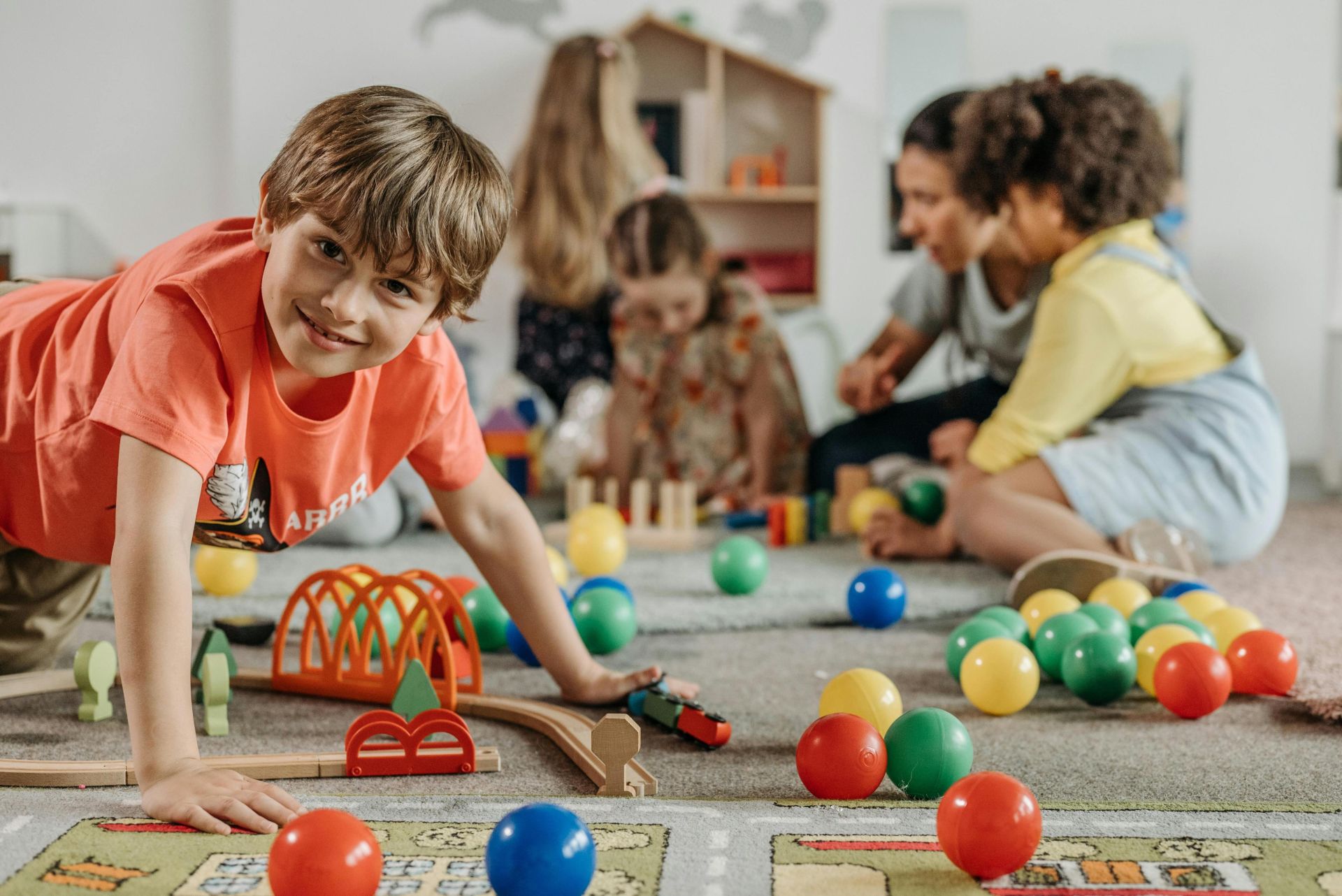 Happy children and a caregiver playing with colorful toys in a lively indoor playroom setting.